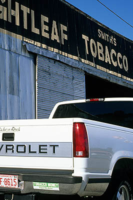 A white Chevy pickup truck in front of Smith's Brighleaf Tobacco Warehouse displays the bumper sticker, "Tobacco Pays My Bills". Location: NC, Pitt County, Farmville, Tobacco warehouses. [ref. to #230.168]