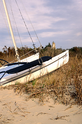 Catamaran beached on dunes; Bald Head Lighthouse (Old Baldy) in bkgd, ctr. Location: NC, Brunswick County, Cape Fear Area, Bald Head Island, West Beach. [ref. to #233.056]