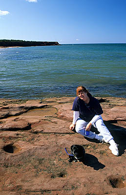 PE: Queens County, Anne's Land Area, Prince Edward Island National Park, Cavendish Beach East. Woman and dog on a red sandstone shelf at the water's edge [Ask for #240.089.]