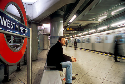 The Underground, Westminster Station. View of the platform; man sits on bench by sign, as train rushes by. NR. Location: ENG, Greater London , City of Westminster, St James District. [ref. to #256.106]