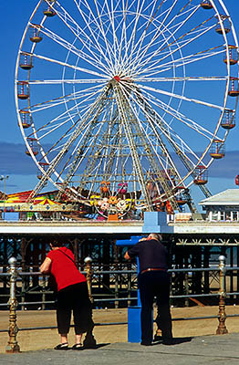 ENG: Manchester-Liverpool Urban Area, Blackpool Borough, The Golden Mile, Central Pier, View past Victorian iron rails along top of seawall, across low tide sands, to Ferris wheel on pier; a couple looks through pay telescope [Ask for #262.020.]